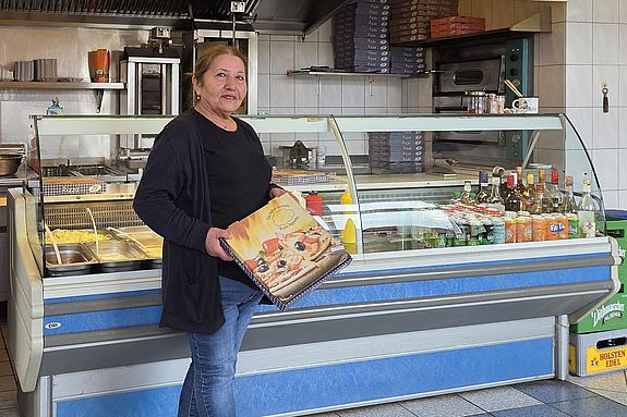 Krümel (Frau Ural) mit einem Pizzakarton in ihrem Grillhaus "Krümel's Grill"- ihre Pizza ist nicht nur im Ort berühmt und beliebt