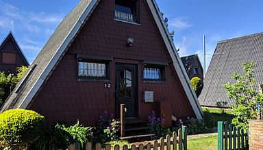 Frontseite des Nurdach-Hauses mit Rasen, grünen Büschen und Gartenzaun vor dem Haus