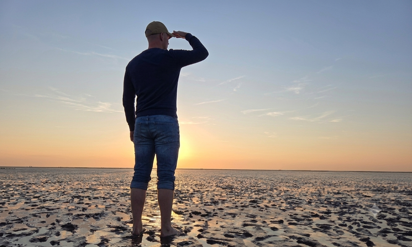 Mann mit Suchpose blickt über das Watt in Richtung Sonnenuntergang